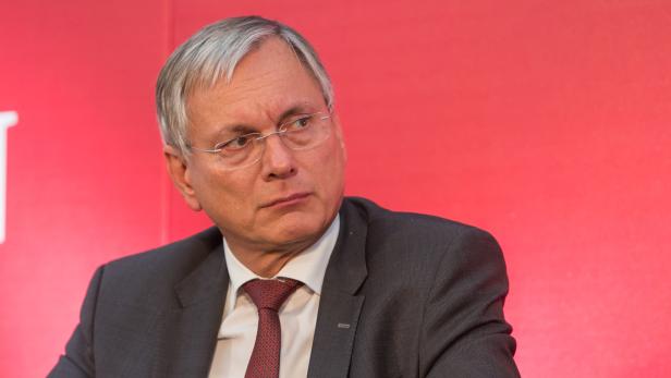 Ein Mann mit Brille, Anzug und Krawatte vor einem roten Hintergrund.