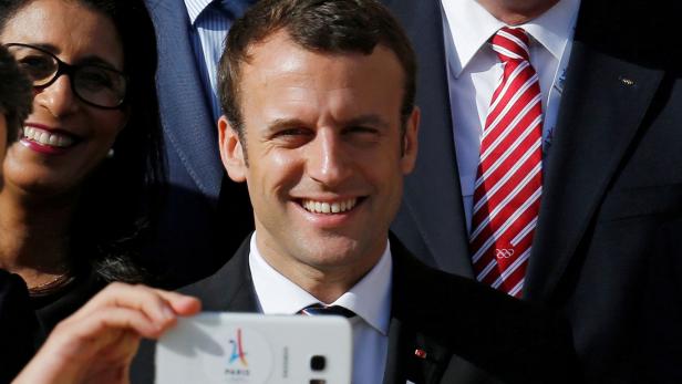 Emmanuel Macron macht ein Selfie mit einem Smartphone, auf dem das Logo von Paris 2024 zu sehen ist.
