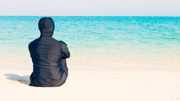 Eine Frau in Burqini sitzt am Strand und blickt auf das Meer.