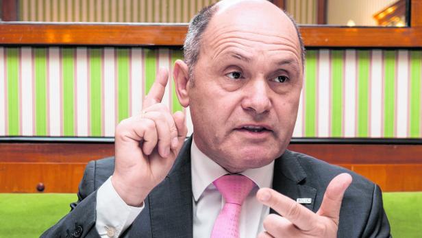 Ein Mann in Anzug und Krawatte gestikuliert vor einem gestreiften Hintergrund.