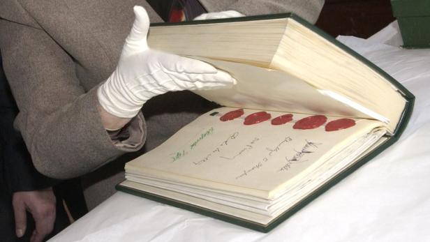 Eine Person mit Handschuhen blättert in einem großen Buch mit Unterschriften und Siegeln.