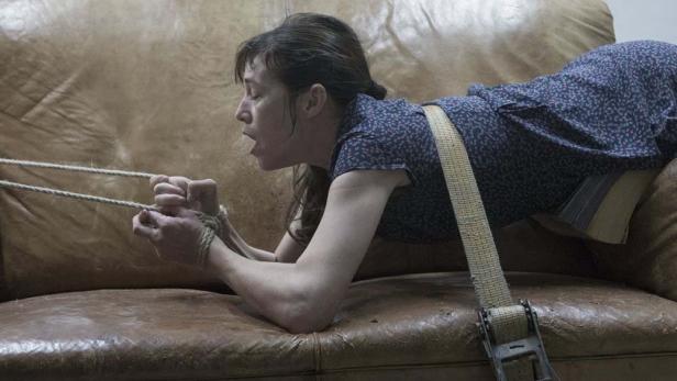 Eine gefesselte Frau liegt auf einem Ledersofa und zieht an einem Seil.