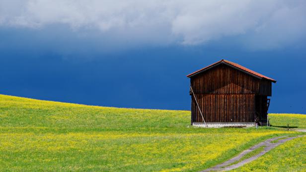 Eine hölzerne Scheune steht auf einer grünen Wiese unter einem dunklen Himmel.