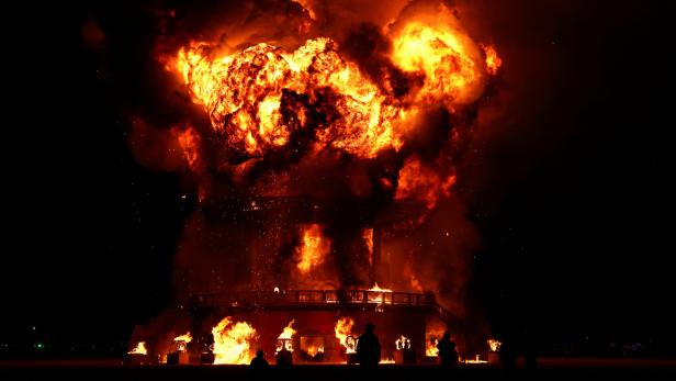 Ein großes Holzgebäude steht in Flammen, umgeben von Rauch und Zuschauern.