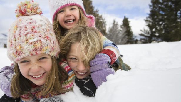 Drei lachende Mädchen liegen im Schnee, gekleidet in Winterkleidung.