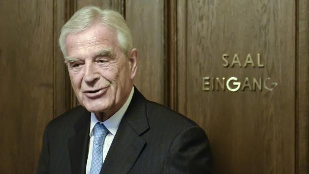 Ein älterer Mann im Anzug steht vor einer Tür mit der Aufschrift „Saal Eingang“.