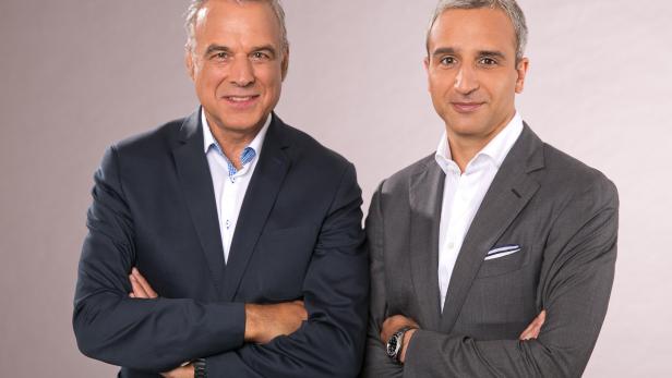 Zwei Männer in Anzügen stehen mit verschränkten Armen vor einem grauen Hintergrund.