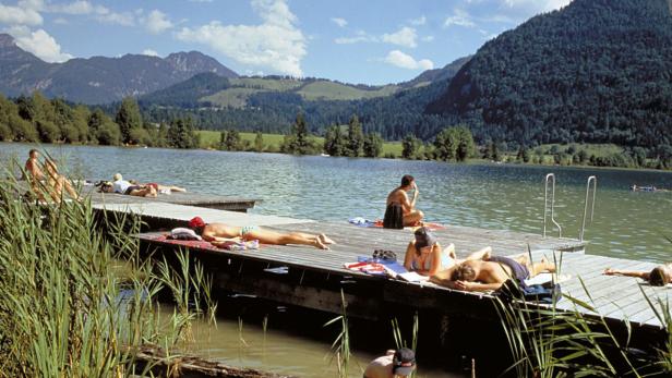 Menschen entspannen sich auf einem Holzsteg an einem See vor einer Bergkulisse.