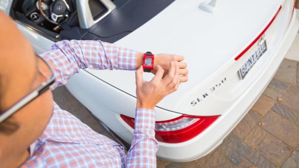 Ein Mann mit Brille betrachtet seine rote Smartwatch vor einem weißen Mercedes-Benz SLK 350.