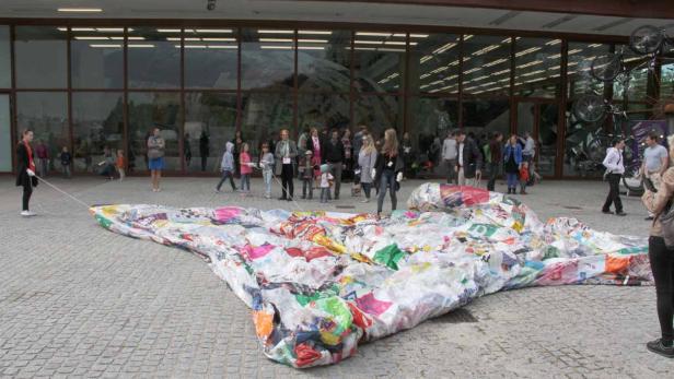 Eine große, bunte Skulptur aus recycelten Plastiktüten vor einem Gebäude mit vielen Menschen im Hintergrund.