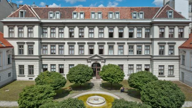 Ein großes, helles Gebäude mit einem kleinen Park und einem Brunnen im Vordergrund.