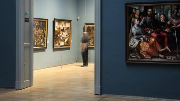 Ein Gemälde hängt in einem Museum, im Hintergrund ein Besucher.