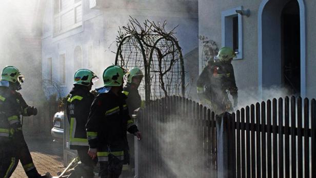 Mehrere Feuerwehrleute stehen vor einem Haus, aus dem Rauch dringt.