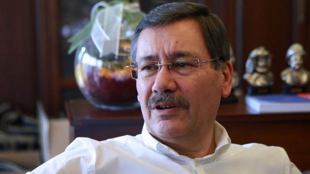 Ein Mann mit Brille und Schnurrbart in einem hellen Hemd sitzt in einem Raum.