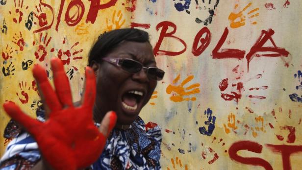 Eine Frau mit erhobener, rot bemalter Hand steht vor einer Wand mit der Aufschrift „Stop Ebola“.