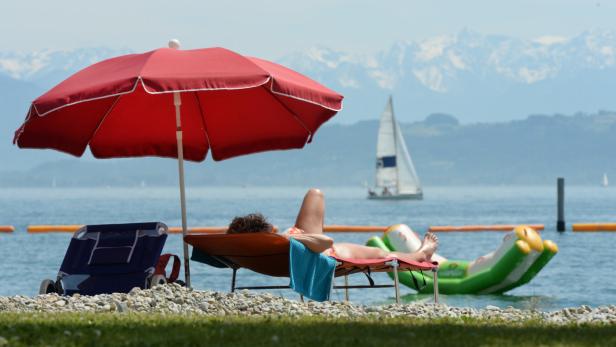 Ein Badegast liegt am 06.06.2014 im Strandbad von Kressbronn (Baden-Württemberg) am Ufer des Bodensees auf einer Liege unter einem Sonnenschirm, während im Hintergrund auf dem Bodensee ein Boot segelt. Foto: Felix Kästle/dpa +++(c) dpa - Bildfunk+++