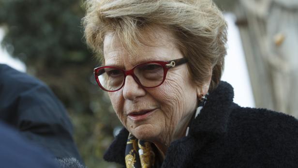 Eine ältere Frau mit roten Brillengläsern und einem dunklen Mantel schaut aufmerksam zu.