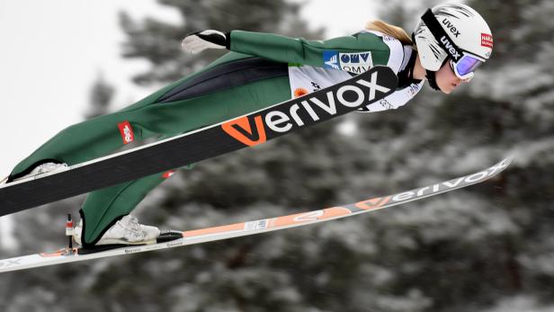 Eine Skispringerin in grüner Sportbekleidung während eines Sprungs.