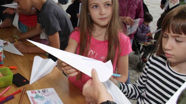 Kinder basteln Papierflieger an einem Tisch mit Stiften und Flyern.