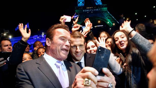 TOPSHOT - Former Governor of California and US actor Arnold Schwarzenegger (L) and French President Emmanuel Macron (C) take a selfie with youths in front of the Eiffel Tower illuminated in green, aboard a boat cruising on the river Seine, after the One Planet Summit in Paris on December 12, 2017. The French President hosts 50 world leaders for the &quot;One Planet Summit&quot;, hoping to jump-start the transition to a greener economy two years after the historic Paris agreement to limit climate change. / AFP PHOTO / POOL / Thibault Camus