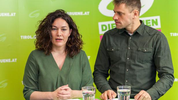Eine Frau und ein Mann sitzen vor einem grünen Hintergrund mit dem Logo der Grünen Tirol.