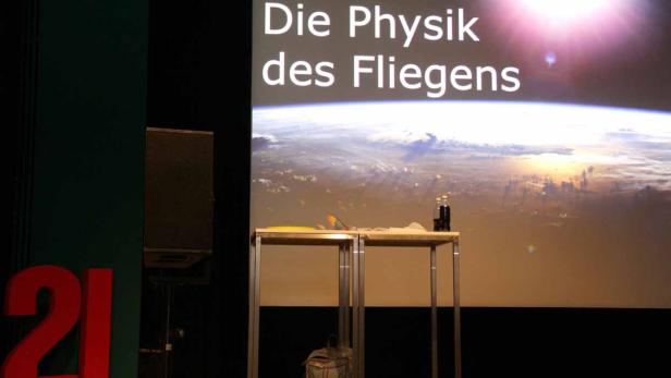 Eine Bühne mit der Aufschrift „Die Physik des Fliegens“ und einem Erdbild im Hintergrund.