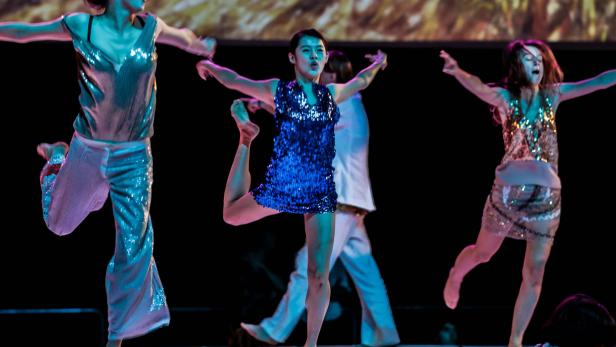 Tänzerinnen in glitzernden Kostümen führen eine Choreografie auf einer Bühne auf.