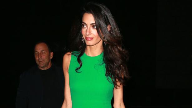 Amal Clooney in einem grünen Kleid, mit einem Mann im Hintergrund.