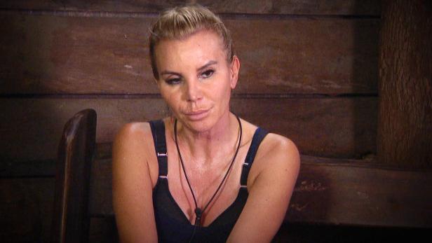 Eine Frau mit blonden Haaren und einem schwarzen Top sitzt vor einer Holzwand.