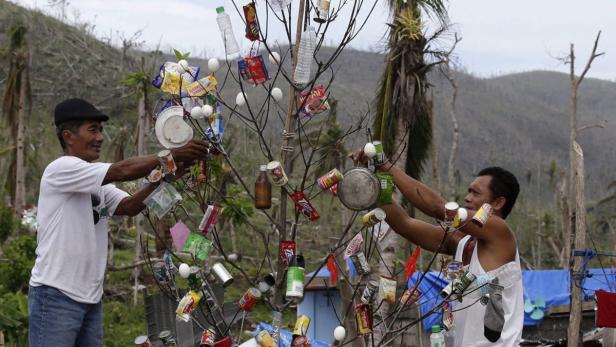 Zwei Männer schmücken einen Baum mit Müll und Essensresten in einer zerstörten Landschaft.