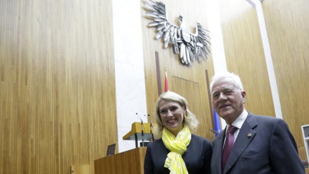 Eine Frau mit gelbem Schal und ein Mann im Anzug vor dem österreichischen Bundesadler.