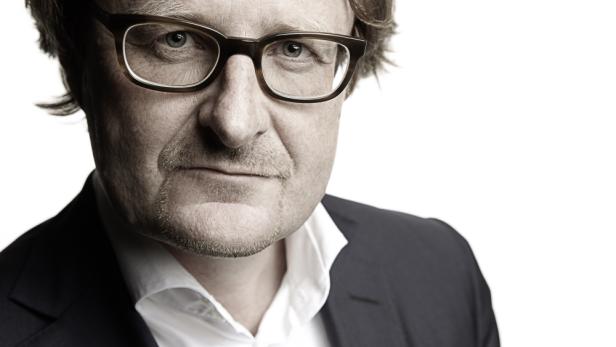 Porträt eines Mannes mit Brille und dunklem Anzug vor einem hellen Hintergrund.