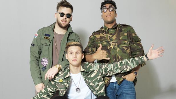 Drei junge Männer posieren in Camouflage-Jacken und mit Schmuck vor einem grauen Hintergrund.