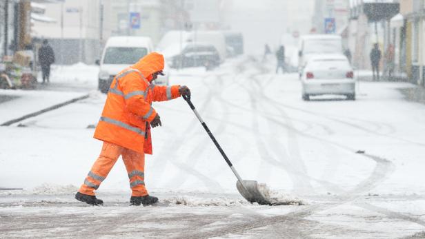 Ein Arbeiter in orangefarbener Kleidung schaufelt Schnee von einer Straße.