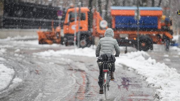 Eine Person fährt bei Schneefall mit dem Fahrrad an Schneepflügen vorbei.