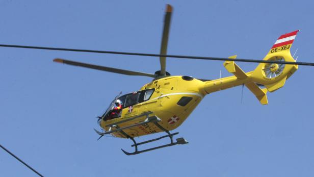 Ein gelber Rettungshubschrauber mit österreichischer Flagge am Heck fliegt vor blauem Himmel.