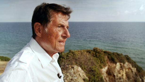Ein Mann in einem weißen Hemd steht vor einer Küstenlandschaft.