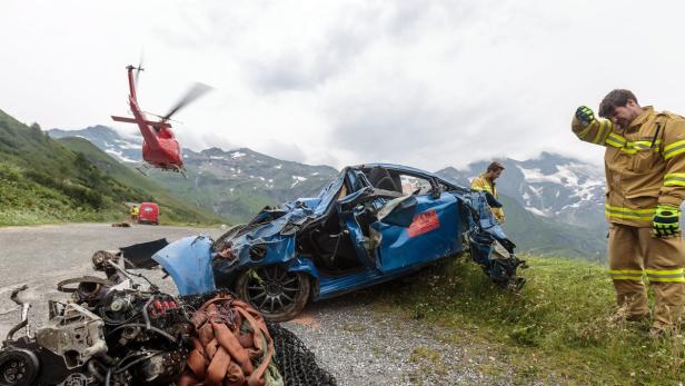 Ein schwer beschädigtes blaues Auto nach einem Unfall in den Bergen, mit Rettungskräften und einem Hubschrauber vor Ort.