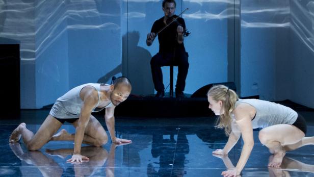 Zwei Tänzer bewegen sich auf einer Bühne, während ein Geiger im Hintergrund sitzt.