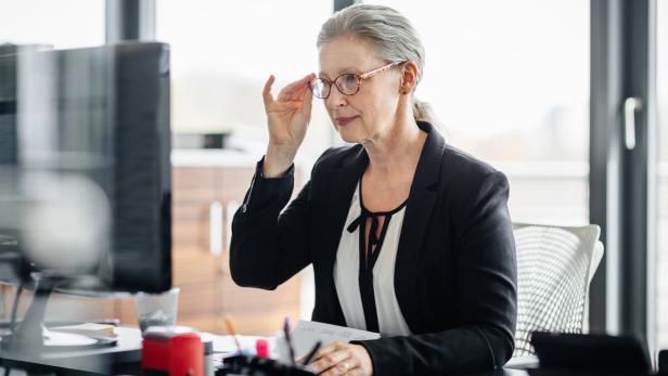 Portrait of a senior businesswoman sitting on her desk in a modern office and. She&#039;s working on a computer and wearing glasses and has silver hair.