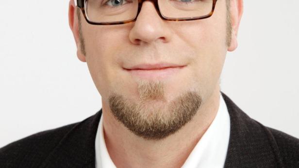 Ein Mann mit Brille, Bart und Anzug vor einem weißen Hintergrund.