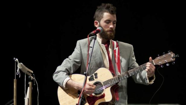 Ein bärtiger Mann mit Anzug spielt Gitarre und singt in ein Mikrofon.