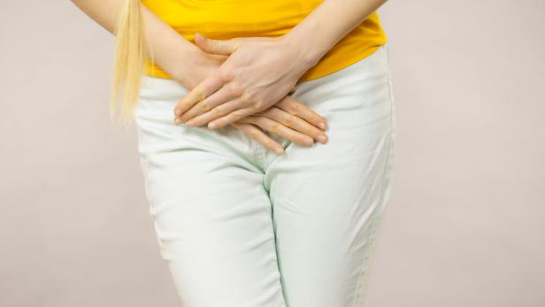 Eine Person hält sich mit beiden Händen den Unterbauch und trägt ein gelbes Oberteil sowie eine helle Hose.