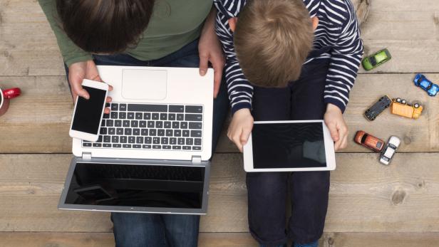 Eine Mutter und ihr Sohn sitzen mit Handy, Laptop und Tablet am Boden