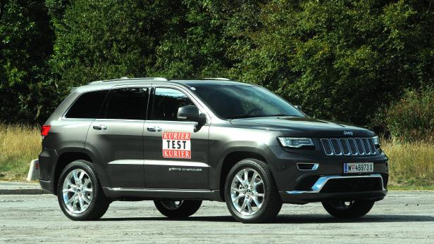 Ein grauer Jeep Grand Cherokee steht vor einer grünen Baumreihe.