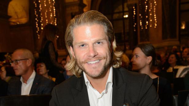 Ein lächelnder Paul Janke mit blonden Haaren sitzt im Anzug in einem Saal mit Publikum.