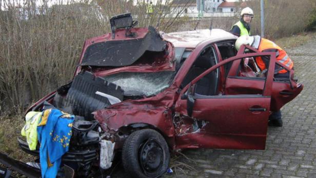 Ein schwer beschädigtes rotes Auto nach einem Unfall, mit Rettungskräften vor Ort.