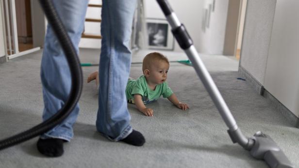 Ein Baby krabbelt auf einem Teppich, während eine Person staubsaugt.