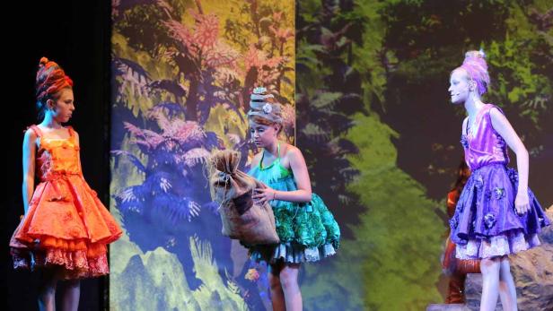 Drei junge Schauspielerinnen in farbenfrohen Kostümen stehen auf einer Bühne vor einer Waldkulisse.