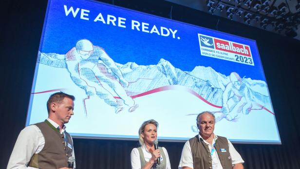 Drei Personen in Tracht stehen vor einer Leinwand mit der Aufschrift „We are ready. Saalbach 2023“.